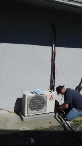 Technician working on an outdoor air conditioning unit against a gray wall.